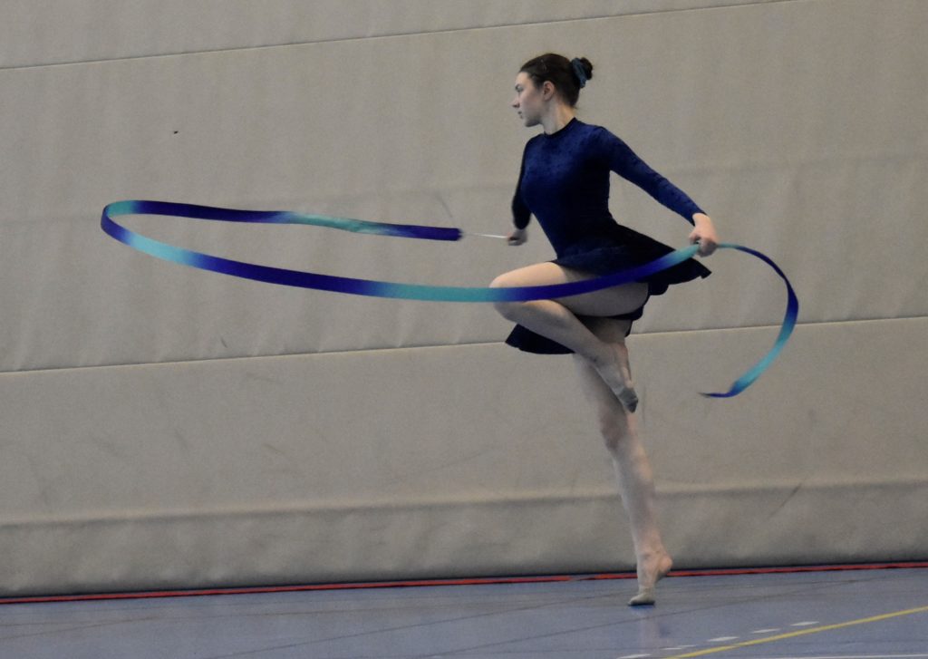 gymnaste en mouvement, sur un pied, tenant un ruban qui forme un demi cercle horizontal autour d'elle